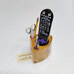 May include: A wooden desk organizer with a curved design, holding a black remote control, a smaller black remote, a yellow pencil, white scissors, and a black marker. The organizer has a natural wood finish.