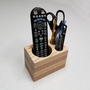 May include: A wooden desk organizer with three compartments. The organizer is made of light wood and has a natural finish. A black remote control, a pair of scissors, and several pens are in the compartments.