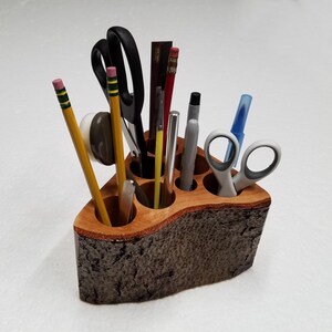 May include: A wooden desk organizer with six compartments holds various writing utensils, including pens, pencils, scissors, and a ruler. The organizer has a dark brown bark-like texture.