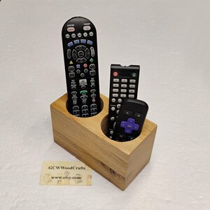 May include: A rectangular light brown wooden remote control holder with two circular cutouts containing three black remote controls. A small rectangular card is placed nearby with the text GCWWoodCrafts and www.etsy.com printed on it.