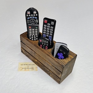 May include: A wooden remote control organizer with a dark brown finish. The organizer has two circular compartments and holds several black remote controls. The organizer is made of wooden planks and has a rectangular shape.