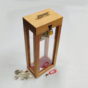 May include: A wooden donation box with a glass front and a lock. The box has a slot on the top for inserting money. A dollar bill is visible inside the box. The box is sitting on a white surface.