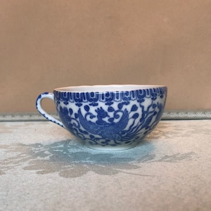 May include: A small, white teacup with a blue and white floral pattern. The cup has a rounded shape and a handle.