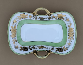 May include: A white porcelain serving dish with two handles. The dish has a green and gold floral design.