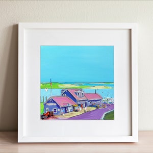 May include: A framed painting of a coastal scene. The artwork depicts blue buildings with pink roofs, a purple road, and a bright blue sky. A red truck is parked in front of the buildings. The painting is in a white frame.
