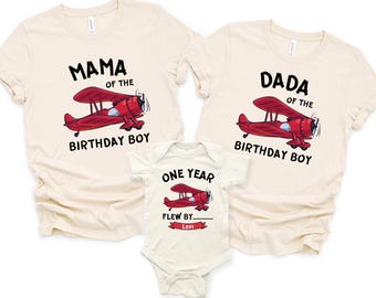 Könnte beinhalten: Ein Set aus cremefarbenen T-Shirts und einem Body. Die Shirts tragen den Text "MAMA of the BIRTHDAY BOY" und "DADA of the BIRTHDAY BOY" mit einer roten Doppeldecker-Grafik. Der Body trägt "ONE YEAR FLEW BY... LEVI" mit einem passenden Doppeldecker.