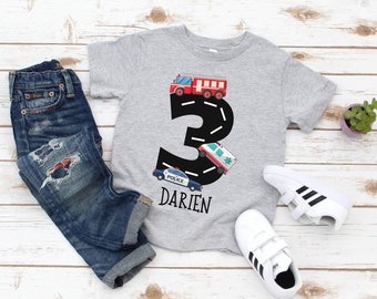 May include: A gray t-shirt with a black number 3 on it. The number 3 is made up of a road with a fire truck, ambulance, and police car driving on it. The text "Darien" is below the number 3.