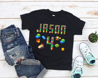 May include: Black t-shirt with a colorful Lego design that says "Jason 4". The shirt is on a white wooden surface with blue jeans, a plant, and two pairs of light blue sneakers.