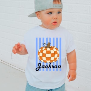 May include: A white short-sleeved t-shirt featuring a graphic of an orange and white checkered pumpkin centered over vertical light blue stripes. The name Jackson is printed in black cursive script below the pumpkin graphic on the front of the shirt.