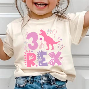 May include: Cream-colored t-shirt with a pink and purple dinosaur-themed graphic. The design includes the number "3", the word "REX", a dinosaur silhouette, a birthday cake, and other festive elements.