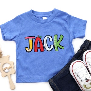 以下が含まれることがあります： 大きなカラフルな文字で「JACK」とプリントされた青い半袖Tシャツ。Jは鉛筆の形、Aは赤いリンゴ、Cは罫線入りのノート用紙のようなデザイン、Kは定規の模様が入った緑色の文字になっています。