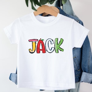 以下が含まれることがあります： 大きなカラフルな文字で「JACK」とプリントされた白い半袖Tシャツ。Jは黄色い鉛筆、Aは赤いリンゴ、Cは罫線入りのノート用紙、Kは目盛りのついた緑色の定規を模したデザインになっています。