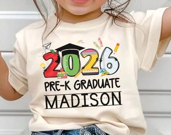 May include: Cream-colored t-shirt with a colorful graphic design. The design features the numbers "2026" with a graduation cap, pencils, and the text "PRE-K GRADUATE MADISON". The shirt is made of a soft material.