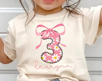May include: Cream-colored t-shirt featuring a pink number 3 decorated with white daisies and a pink bow. The name "laurynn" is written in cursive below the number. The shirt is worn by a child.