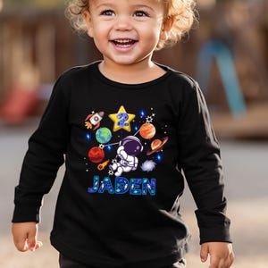 May include: Black long-sleeve shirt with a colorful space-themed graphic. The design includes a rocket, planets, stars, and an astronaut. The number "2" is in a yellow star, and the name "Jaden" is displayed in blue letters.