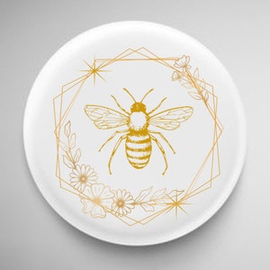 Pode incluir: Um prato de cer&acirc;mica branco com um design geom&eacute;trico dourado que apresenta uma abelha e detalhes florais.