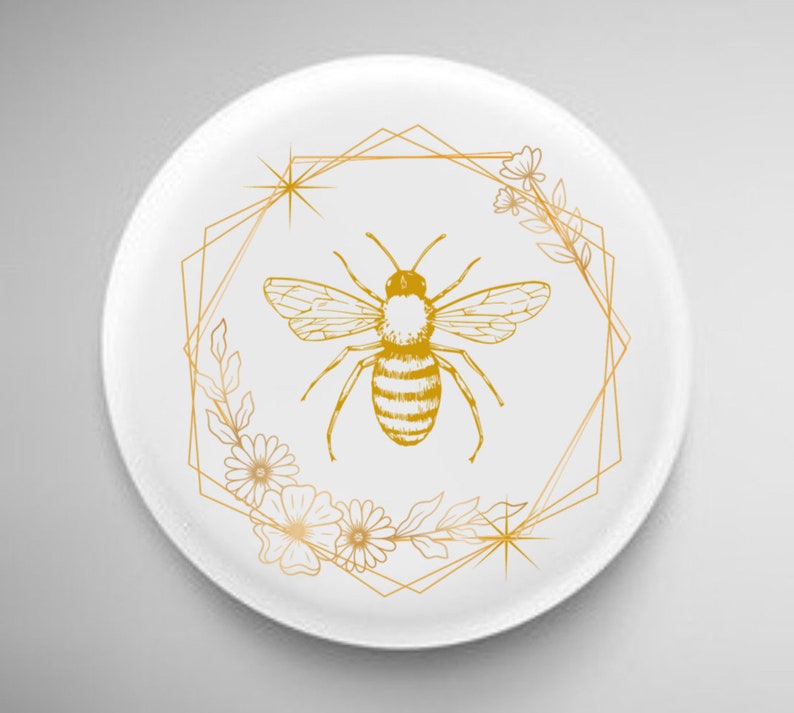 Pode incluir: Um prato de cer&acirc;mica branco com um design geom&eacute;trico dourado que apresenta uma abelha e detalhes florais.