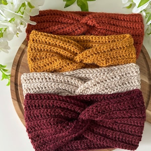 May include: Four crocheted headbands in different colours: burgundy, beige, mustard yellow, and burnt orange. The headbands are stacked on top of each other on a wooden surface.
