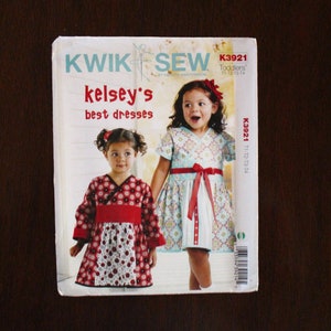 May include: Kwik Sew sewing pattern for two girls' dresses. The pattern is called "Kelsey's Best Dresses" and includes two designs, one with a red and white floral print and the other with a blue and white floral print. The pattern is for toddlers sizes 11-12, 13-14. The pattern number is K3921.
