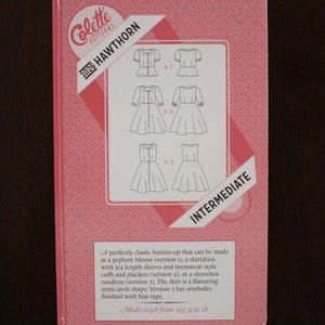 May include: Pink sewing pattern package for the Colette Hawthorne dress. The package features illustrations of the dress variations, including a peplum blouse and a sleeveless sundress. The difficulty level is intermediate.