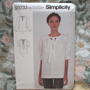 May include: A Simplicity sewing pattern packet, S9233 H5, featuring a woman wearing a white blouse with a tie-neck detail. The packet includes illustrations of the blouse and sizing information.