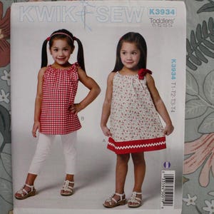 May include: Kwik Sew pattern K3934 for toddlers sizes T1-T4. The envelope shows two outfits: a red and white gingham sleeveless top with white pants, and a cream dress with a small floral print, a red hem, and a red shoulder bow.