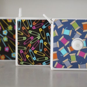 May include: Three white retractable tape measures with different colorful patterns. The first has a black background with colorful flowers and pins. The second has a black background with colorful safety pins. The third has a blue background with colorful spools of thread.