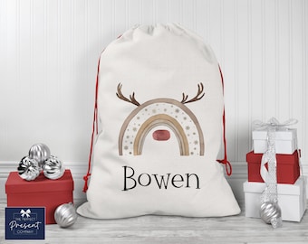 May include: A white personalised Christmas gift bag with a red drawstring. The bag features a watercolour illustration of a rainbow with a reindeer face and the name "Bowen" printed below.