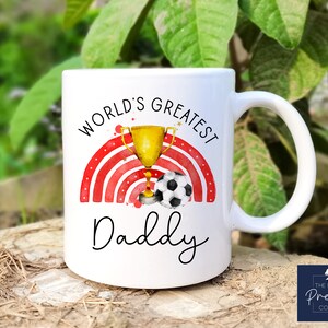 Puede incluir: Taza de cer&aacute;mica blanca con un dise&ntilde;o de arco&iacute;ris rojo y blanco, un trofeo dorado con un bal&oacute;n de f&uacute;tbol y el texto "World's Greatest Daddy".