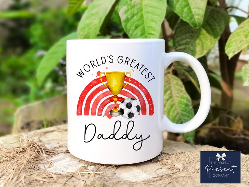 Puede incluir: Taza de cer&aacute;mica blanca con un dise&ntilde;o de arco&iacute;ris rojo y blanco, un trofeo dorado con un bal&oacute;n de f&uacute;tbol y el texto "World's Greatest Daddy".