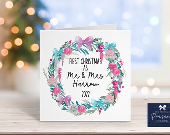 May include: A white Christmas card with a wreath of pink and blue ornaments, greenery, and a pink bow. The text reads "First Christmas as Mr & Mrs Harrow 2022".