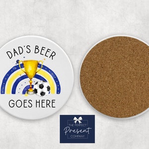 Puede incluir: Posavasos de cer&aacute;mica blanca con un arco&iacute;ris azul y amarillo, un trofeo dorado y un bal&oacute;n de f&uacute;tbol. El texto "Dad's Beer Goes Here" est&aacute; impreso en el posavasos.