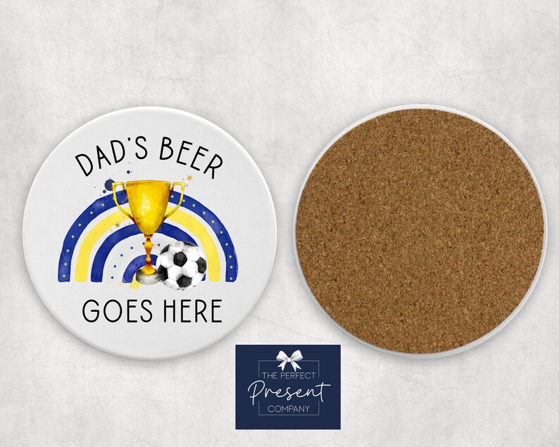 Puede incluir: Posavasos de cer&aacute;mica blanca con un arco&iacute;ris azul y amarillo, un trofeo dorado y un bal&oacute;n de f&uacute;tbol. El texto "Dad's Beer Goes Here" est&aacute; impreso en el posavasos.