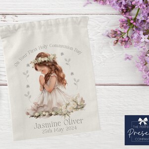 May include: White drawstring bag with a watercolor illustration of a girl praying. The bag has the text "On Your First Holy Communion Day" and "Jasmine Oliver 25th May 2024".
