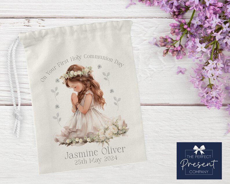 May include: White drawstring bag with a watercolor illustration of a girl praying. The bag has the text "On Your First Holy Communion Day" and "Jasmine Oliver 25th May 2024".