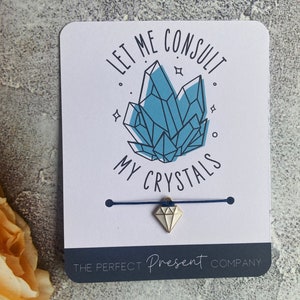 May include: A white card with blue text that reads "Let me consult my crystals." A blue string bracelet with a silver diamond charm is attached to the card. The card also has the text "The Perfect Present Company" in black text.