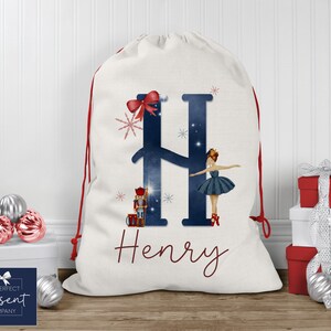 Puede incluir: Una media de Navidad blanca con una letra H azul y blanca, un lazo rojo y una ilustración de bailarina. La media está personalizada con el nombre "Henry".