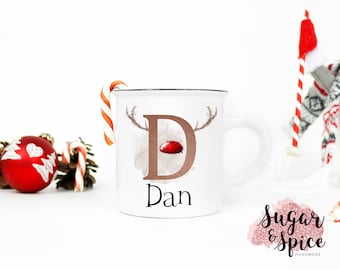 May include: White ceramic mug with a brown letter "D" with a red nose and antlers, the name "Dan" is written below the letter. The mug is next to a candy cane and a pine cone.