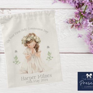 May include: A white drawstring bag with a watercolor illustration of a girl in a white dress with a flower crown, praying. The text "On Your First Holy Communion Day" is above the girl and "Harper Milnes 25th May 2024" is below the girl. The bag is on a white wooden surface.