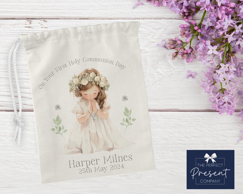 May include: A white drawstring bag with a watercolor illustration of a girl in a white dress with a flower crown, praying. The text "On Your First Holy Communion Day" is above the girl and "Harper Milnes 25th May 2024" is below the girl. The bag is on a white wooden surface.
