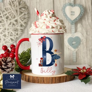 May include: White ceramic mug with a red handle. The mug features a blue letter "B" with a ballerina and a nutcracker design. The name "Billy" is written below the letter. The mug is topped with whipped cream and candy canes.