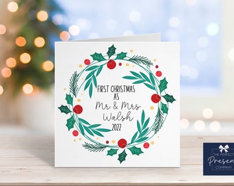 May include: A white Christmas card with a green wreath decorated with red berries and gold dots. The text on the card reads "First Christmas as Mr & Mrs Walsh 2022".