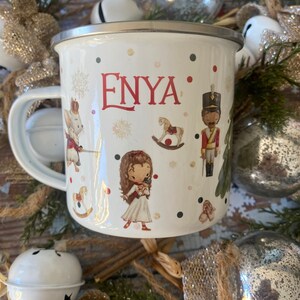 Pode incluir: Caneca de esmalte branco com um design de floco de neve vermelho e branco. A caneca tem uma borda e uma alça prateadas. O nome "ENYA" está impresso em vermelho na caneca. A caneca está cercada por enfeites de Natal.