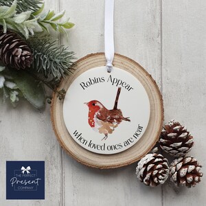 Op de afbeelding: Een ronde, witte kerstversiering met een aquarel illustratie van een roodborstje en de tekst "Robins Appear when loved ones are near". De versiering is bevestigd aan een houten schijf met een wit lint om op te hangen. Pijnappels en groen zijn op de achtergrond.