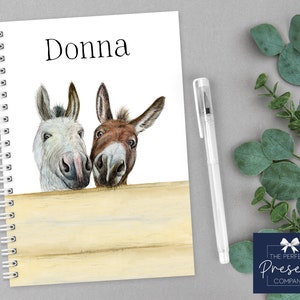 May include: A white spiral bound notebook with the name "Donna" printed on the front cover. The cover features a watercolour illustration of two donkeys peeking over a wooden fence.