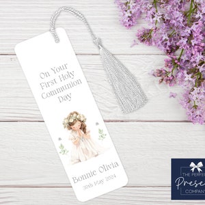 May include: White bookmark with a silver tassel. The bookmark features a watercolor illustration of a girl in a white dress with a flower crown, praying. The text reads "On Your First Holy Communion Day" and "Bonnie Olivia 30th May 2024".