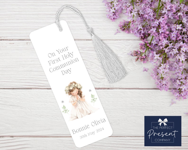 May include: White bookmark with a silver tassel. The bookmark features a watercolor illustration of a girl in a white dress with a flower crown, praying. The text reads "On Your First Holy Communion Day" and "Bonnie Olivia 30th May 2024".