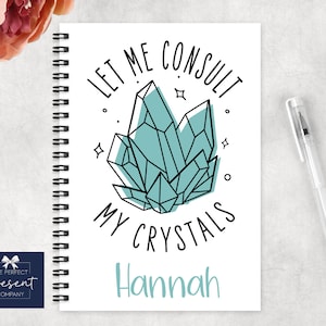 May include: A white spiral bound notebook with the text "Let me consult my crystals" and a drawing of teal blue crystals. The name "Hannah" is written below the crystals.