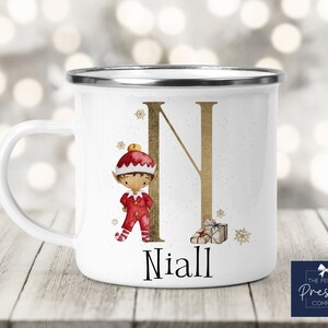 Può includere: Tazza in smalto bianco con una lettera N dorata, un'illustrazione di un elfo di Natale e il nome Niall. La tazza ha un bordo argentato.