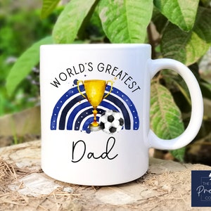 Puede incluir: Taza de cer&aacute;mica blanca con un dise&ntilde;o de arco&iacute;ris azul y blanco, un trofeo dorado y un bal&oacute;n de f&uacute;tbol. El texto "World's Greatest Dad" est&aacute; impreso en la taza.
